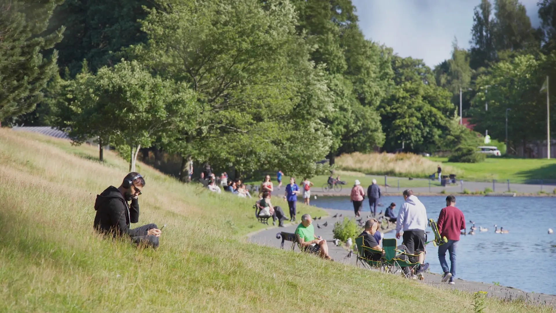 People relaxing and walking beside a tree-lined park lake on a sunny day