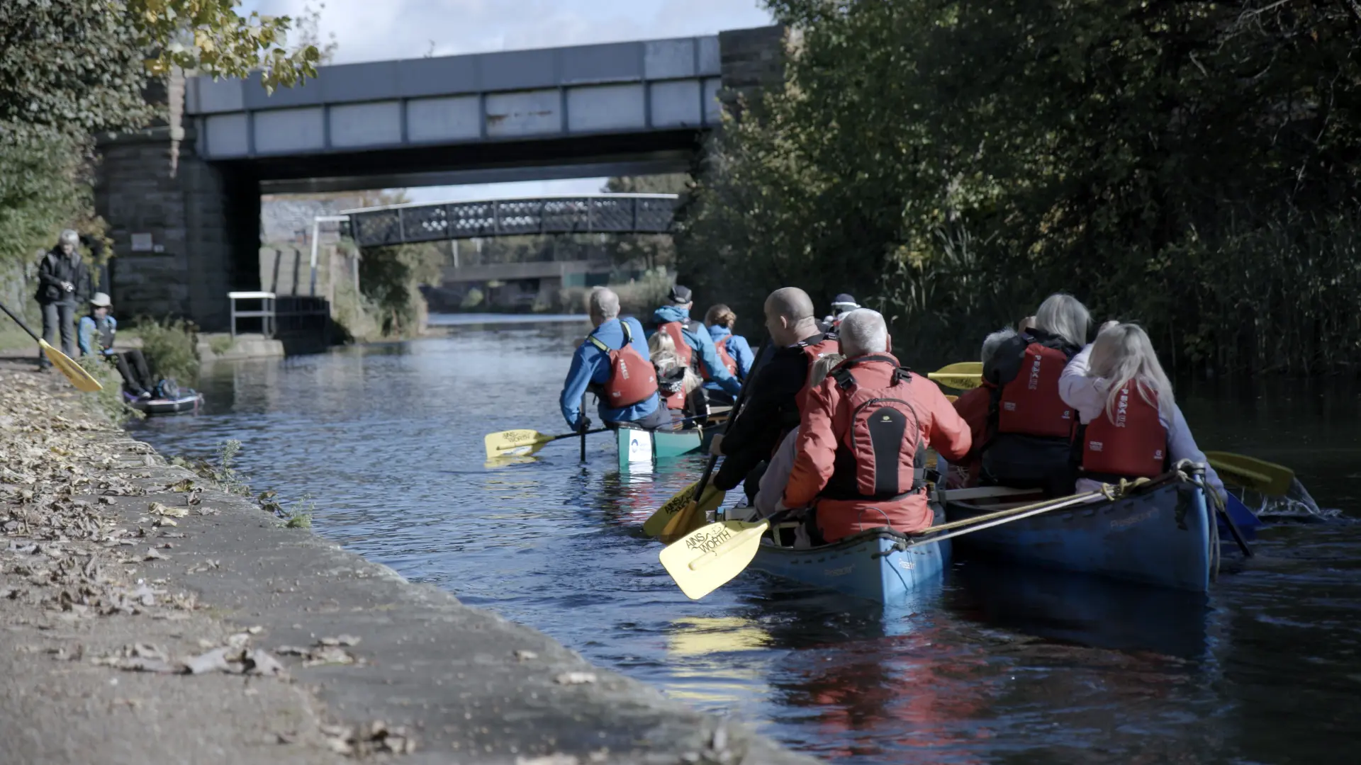 A group of people paddling canoes along an urban canal under a bridge