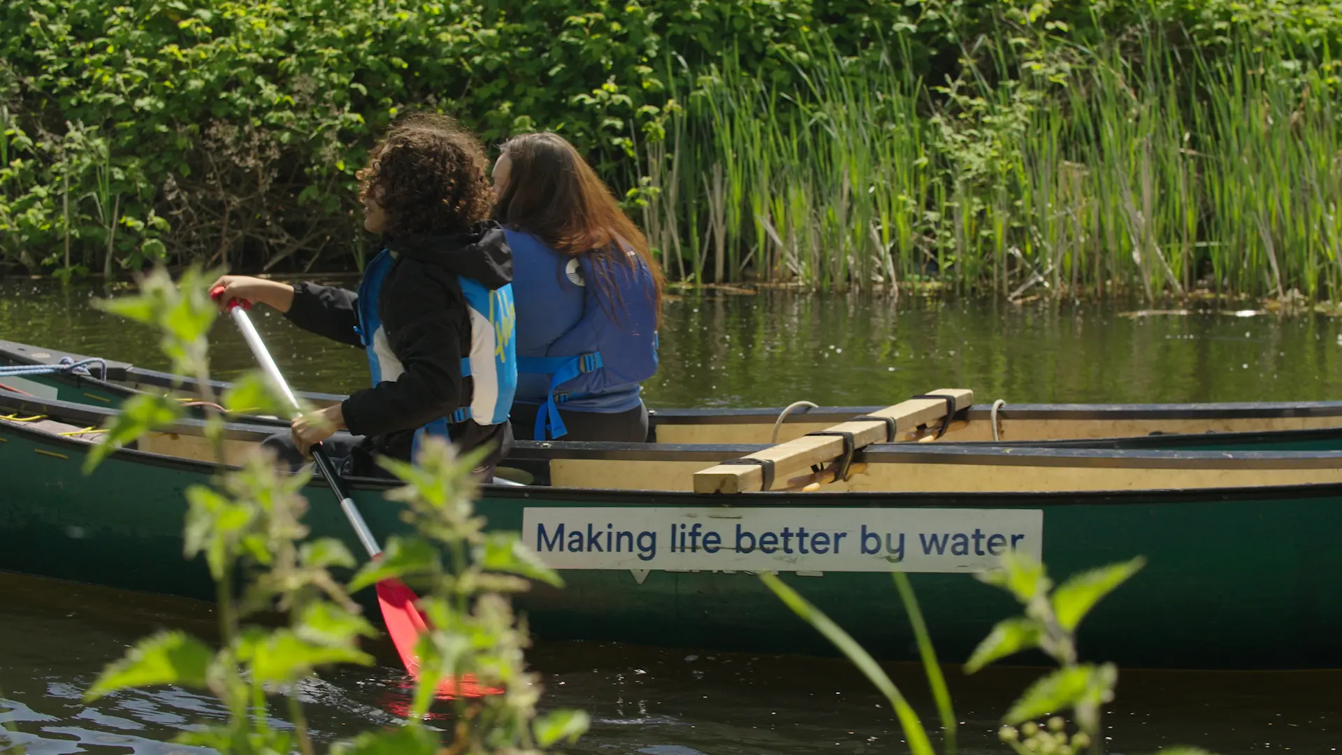 Two people in life jackets paddling a green canoe along a leafy waterway, with 'Making life better by water' written on the hull